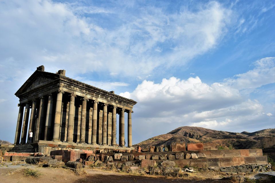 Un tempio classico in pietra con un grande colonnato si erge su una piattaforma di pietra in rovina in un paesaggio collinare e arido sotto un cielo nuvoloso.