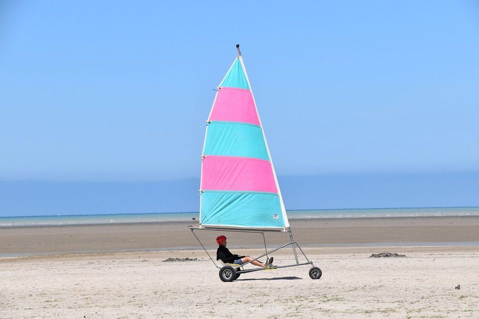 Une personne dans un char à voile avec une voile rose et turquoise est assise sur une large plage de sable près de l'océan sous un ciel bleu clair.