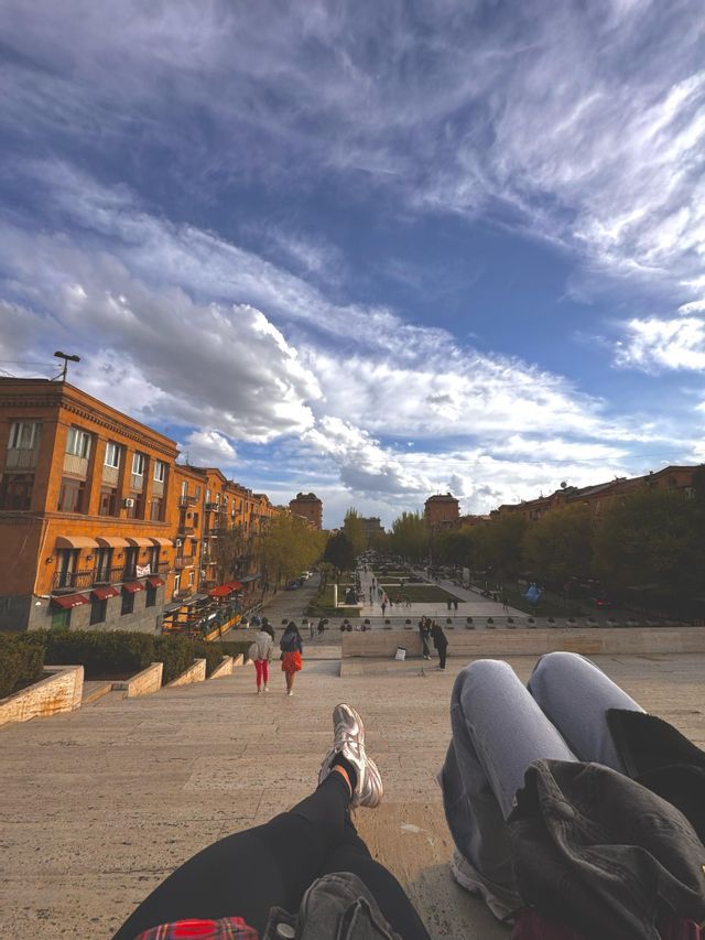 Una vista in prima persona di due persone sedute su un'ampia scalinata in pietra che si affaccia su una piazza cittadina con persone che camminano sotto.
