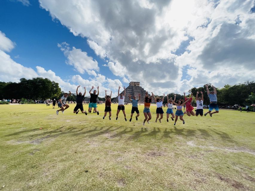 A WeRoad group trip jumps in the air on a green lawn, with a large stone pyramid in the background under a cloudy sky.
