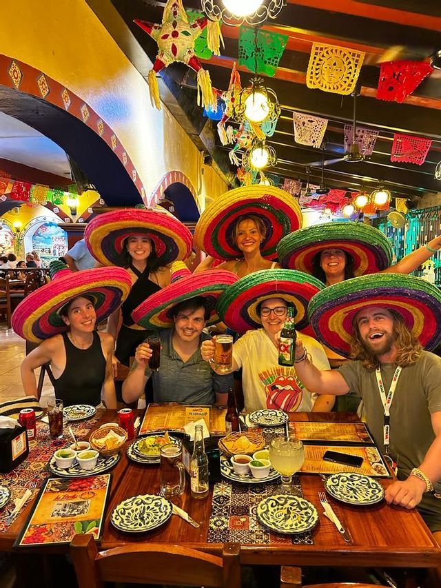 A WeRoad group trip wearing colorful sombreros smiles for a photo while sitting at a table in a decorated restaurant.