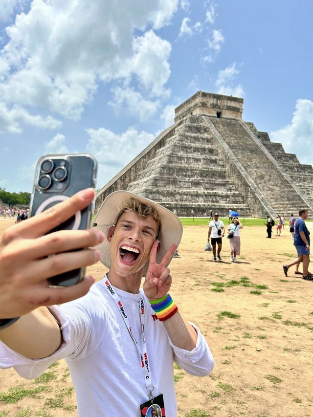 A smiling man on a WeRoad group trip takes a selfie with a peace sign in front of a large ancient pyramid.
