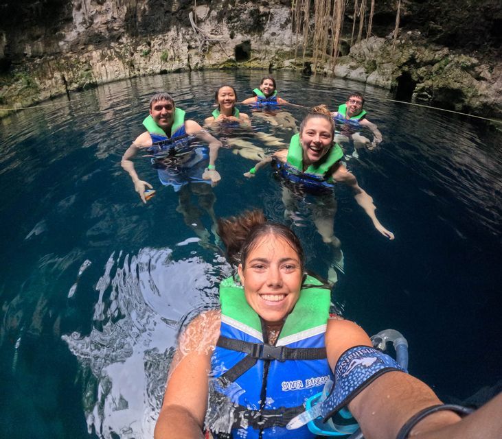 A smiling WeRoad group trip in life vests takes a selfie while swimming in the clear blue water of a cenote.