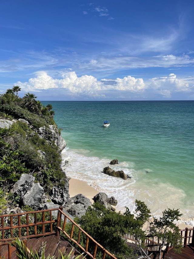 A high-angle view of a small boat on turquoise water, next to a sandy beach and a rocky cliff with palm trees.