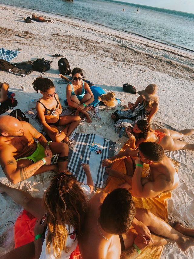 A WeRoad group trip sitting in a circle on a sandy beach, playing cards together as the sun sets over the water.
