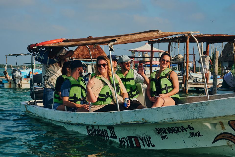A WeRoad group trip wearing green life jackets smiles and waves while sitting in a small boat on the water near a dock.