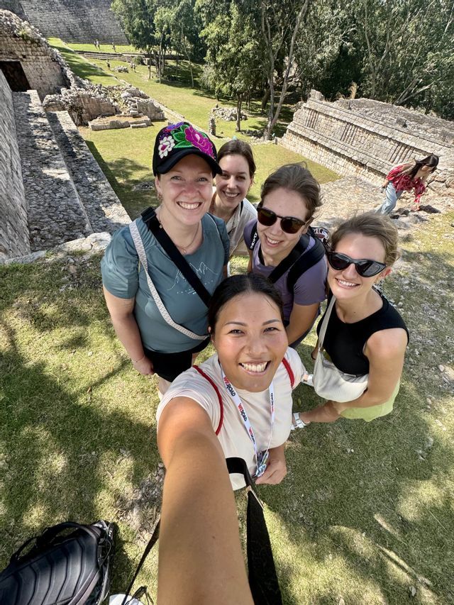 A WeRoad group trip of five women smiling for a selfie in front of ancient stone ruins on a grassy slope.