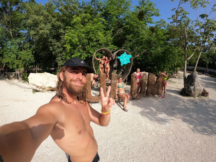 A man takes a selfie with a WeRoad group trip as they pose on and around a large woven sculpture in a park with lush green trees.
