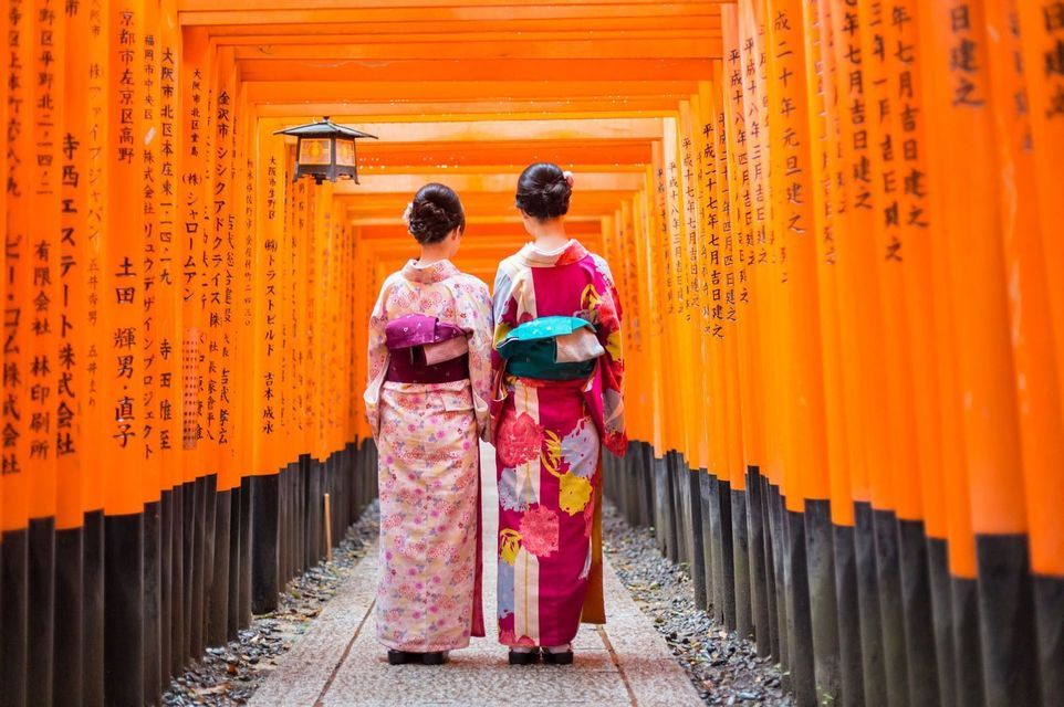 Two women seen from behind in traditional kimonos standing on a path through a tunnel of orange torii gates.