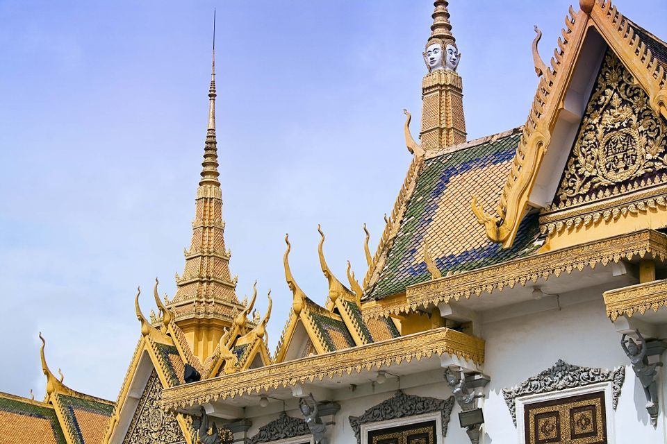 Flèches dorées ornées et toits étagés et colorés d'un temple cambodgien, avec une tour présentant quatre visages sculptés sur un ciel bleu.