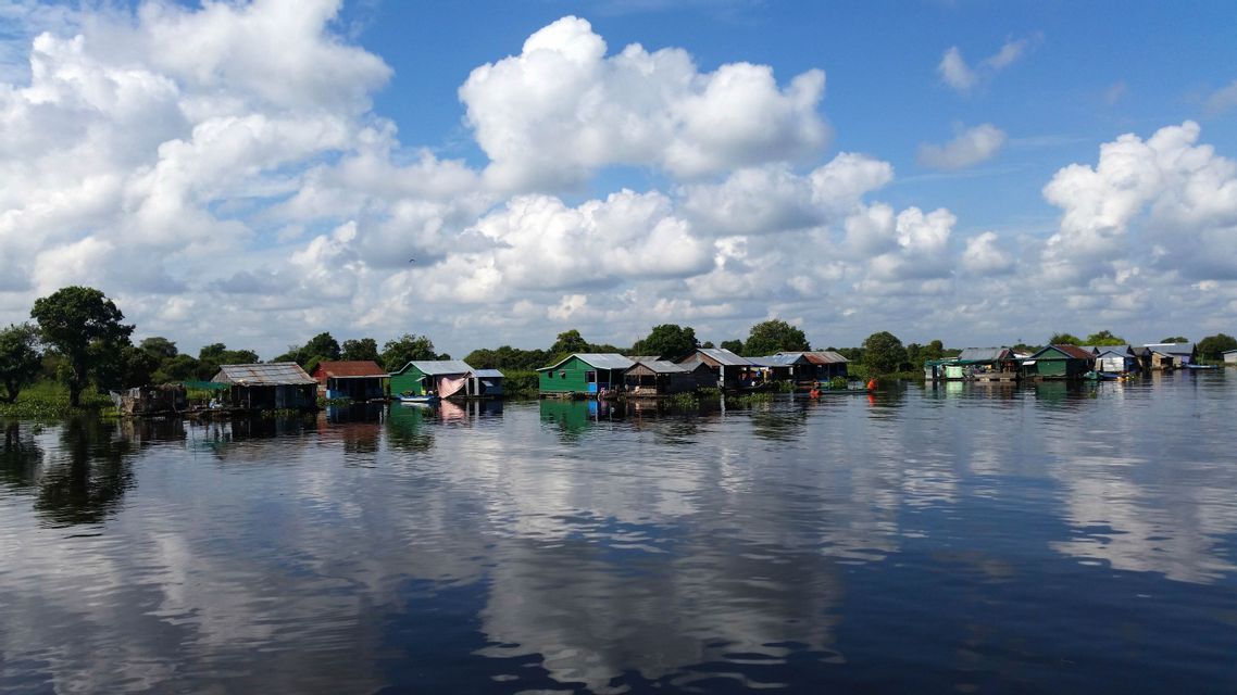 Un village flottant avec des maisons colorées sur une rivière calme, reflétant le ciel bleu et les nuages blancs.