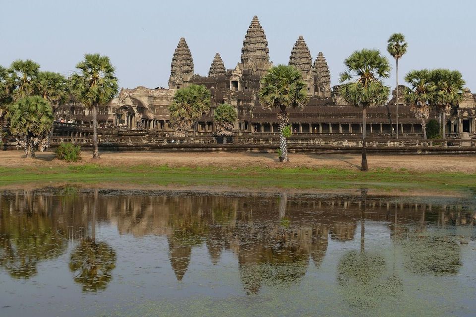 Un ancien temple de pierre avec plusieurs tours et des palmiers se reflète dans une étendue d'eau calme au premier plan.