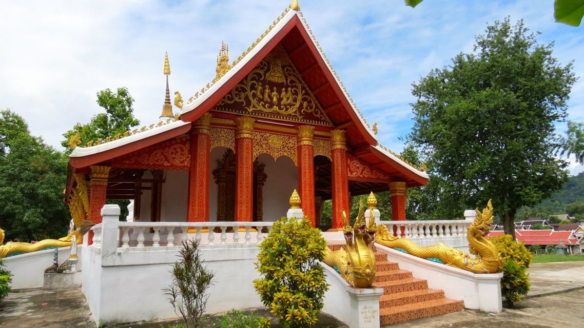 Un temple bouddhiste orné de rouge et d'or, avec des statues de naga dorées le long de son escalier, se dressant sur fond d'arbres et de ciel bleu.