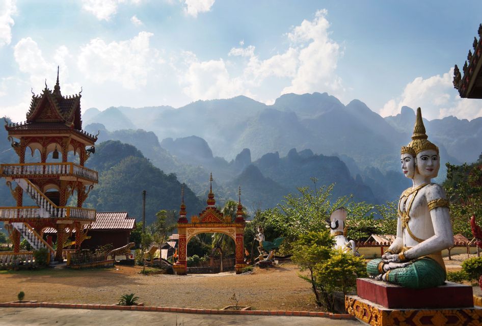 Une statue à plusieurs têtes et une tour ornée dans un complexe de temples avec en toile de fond des montagnes vertes et brumeuses.