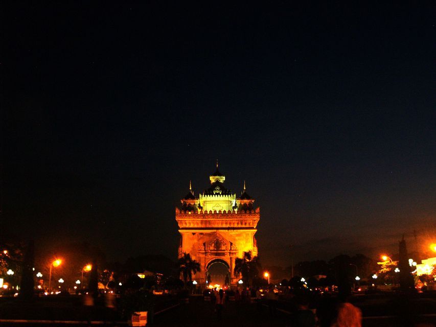 Un grand monument orné illuminé de lumières dorées sur un ciel nocturne sombre, avec des réverbères et des arbres en contrebas.