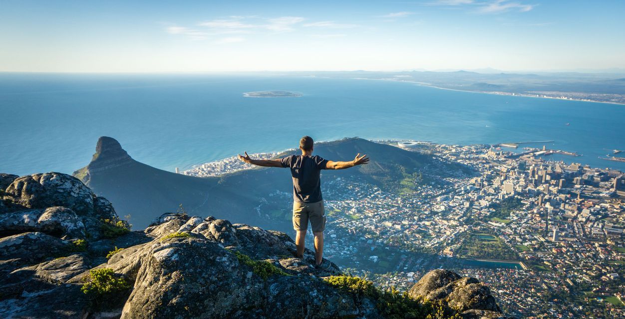 Un homme vu de dos se tient sur un sommet rocheux, les bras tendus, surplombant une vaste ville côtière et la mer.
