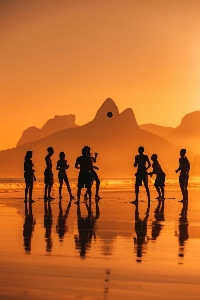 Silhouettes of a WeRoad group trip playing with a ball on a wet beach at sunset, with mountains in the background.
