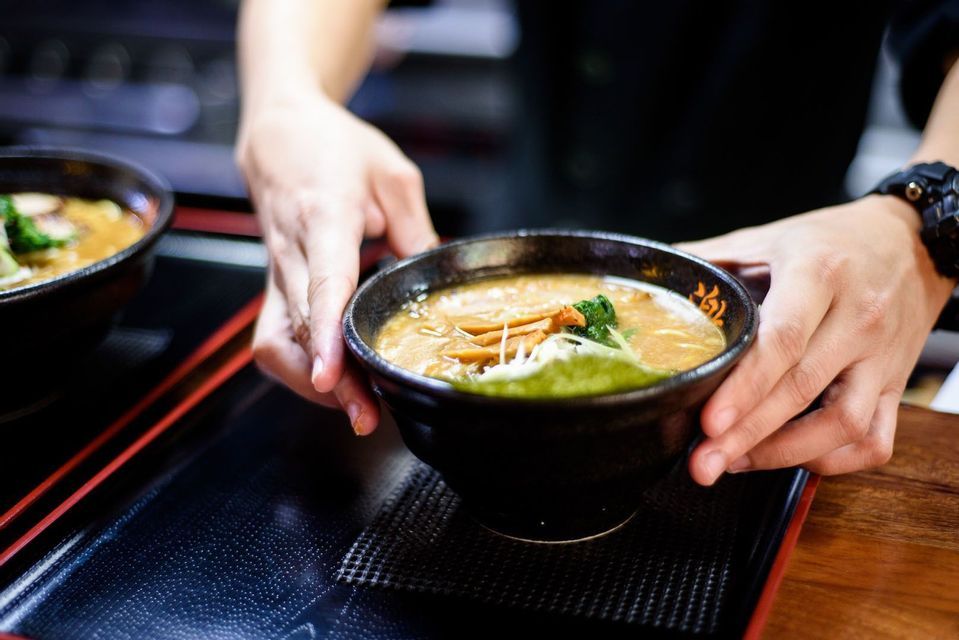 A person serves a bowl of ramen soup with toppings, placing it on a black tray.