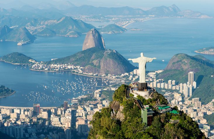 The Christ the Redeemer statue stands on a mountain peak overlooking a coastal city, Sugarloaf Mountain, and a bay filled with boats.