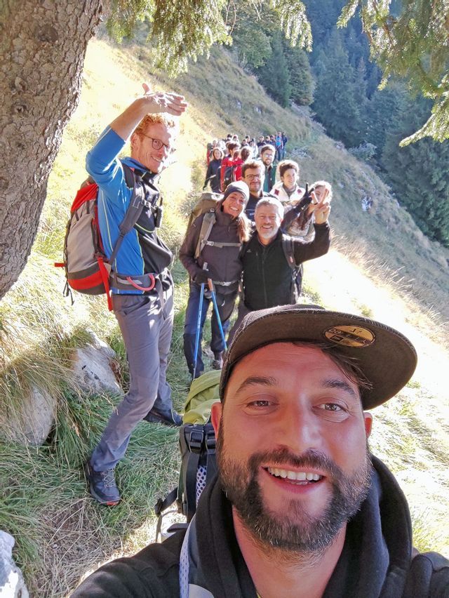 Un uomo si scatta un selfie sorridente con un gruppo WeRoad durante un'escursione in fila indiana su un sentiero di montagna soleggiato.