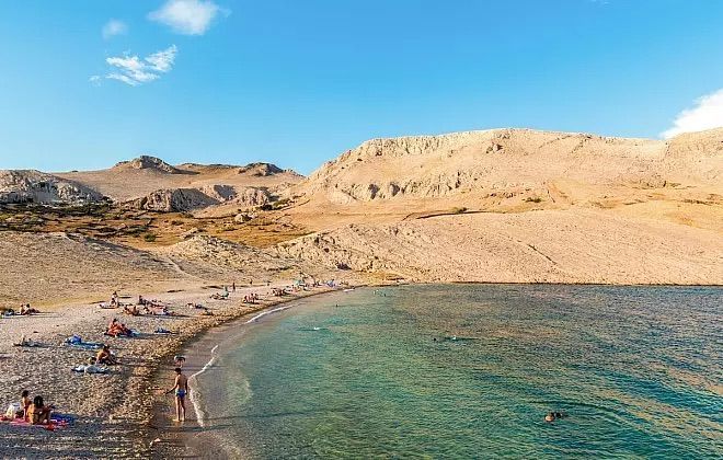 Persone che prendono il sole e nuotano in una caletta, ai piedi di aride colline color sabbia, sotto un cielo azzurro.