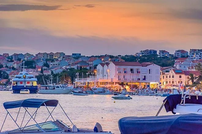 Barche ormeggiate in un porto di fronte a una città costiera arroccata su una collina, illuminata al tramonto.