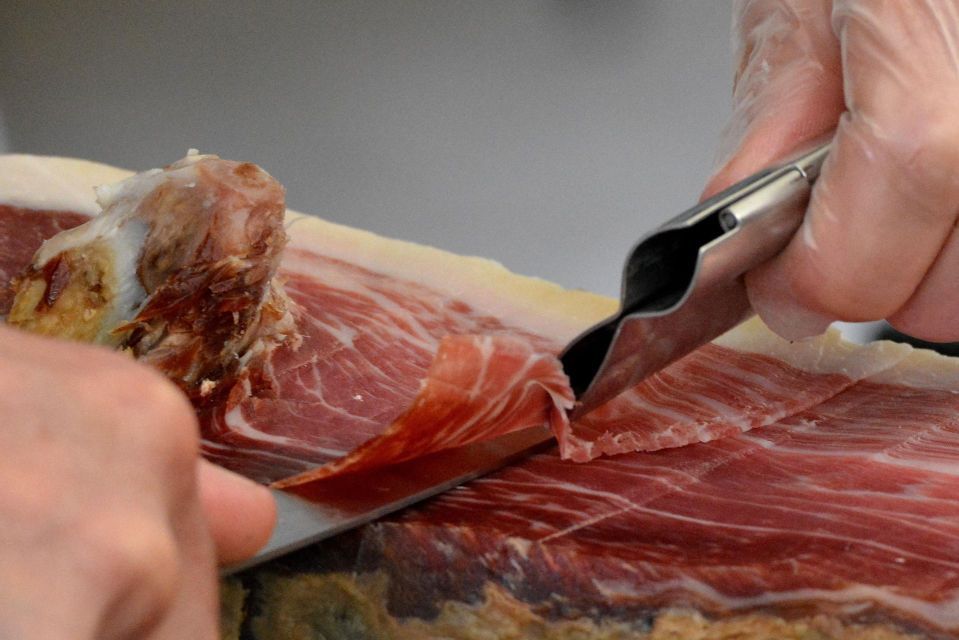 Primer plano de una persona con guante usando pinzas y un cuchillo para cortar una fina loncha de jamón curado.