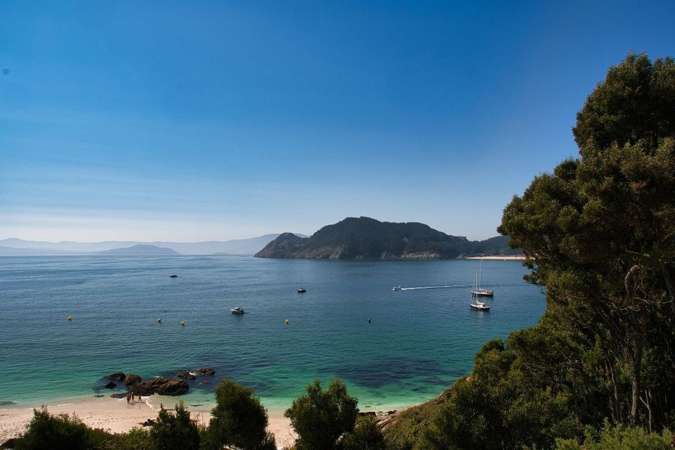 Vista aérea de una bahía con barcos en agua turquesa, una playa de arena blanca y una isla boscosa bajo un cielo despejado.