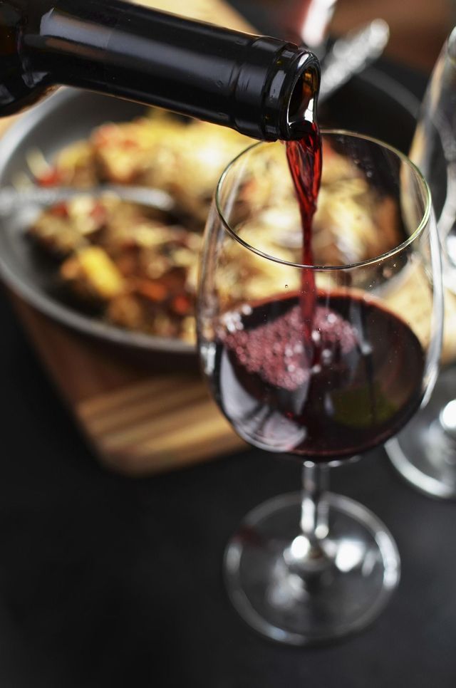 Vino tinto siendo vertido de una botella a una copa, con un plato de comida borroso de fondo.