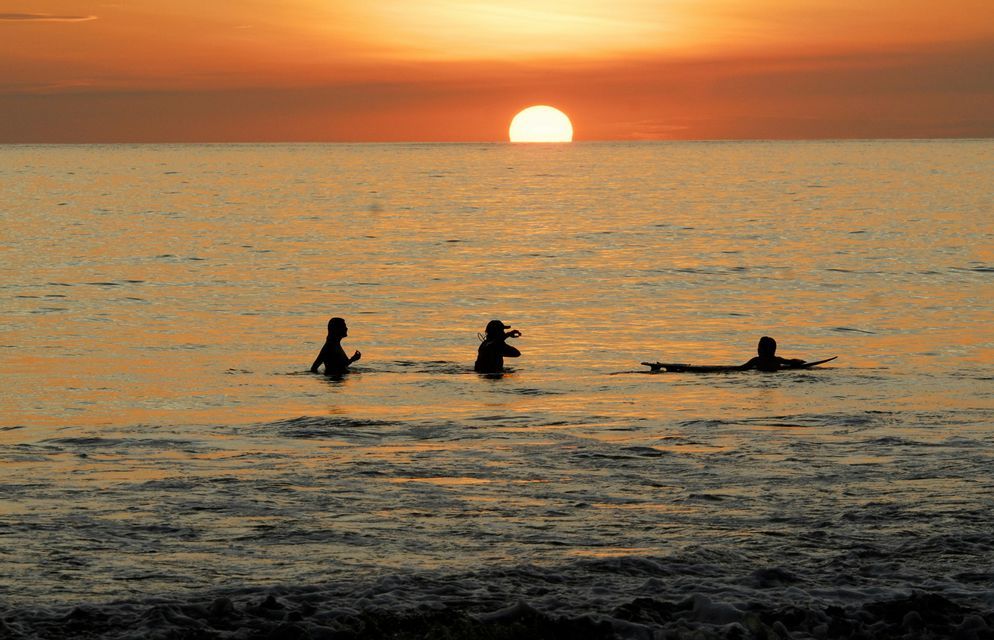 Tres personas de un viaje en grupo de WeRoad silueteadas en el océano durante un atardecer anaranjado, una de ellas en una tabla de surf.