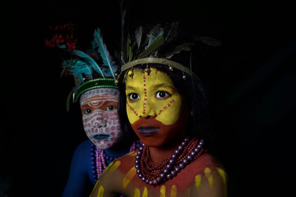 Primer plano de dos jóvenes con elaborada pintura facial tradicional y tocados de plumas sobre fondo negro.