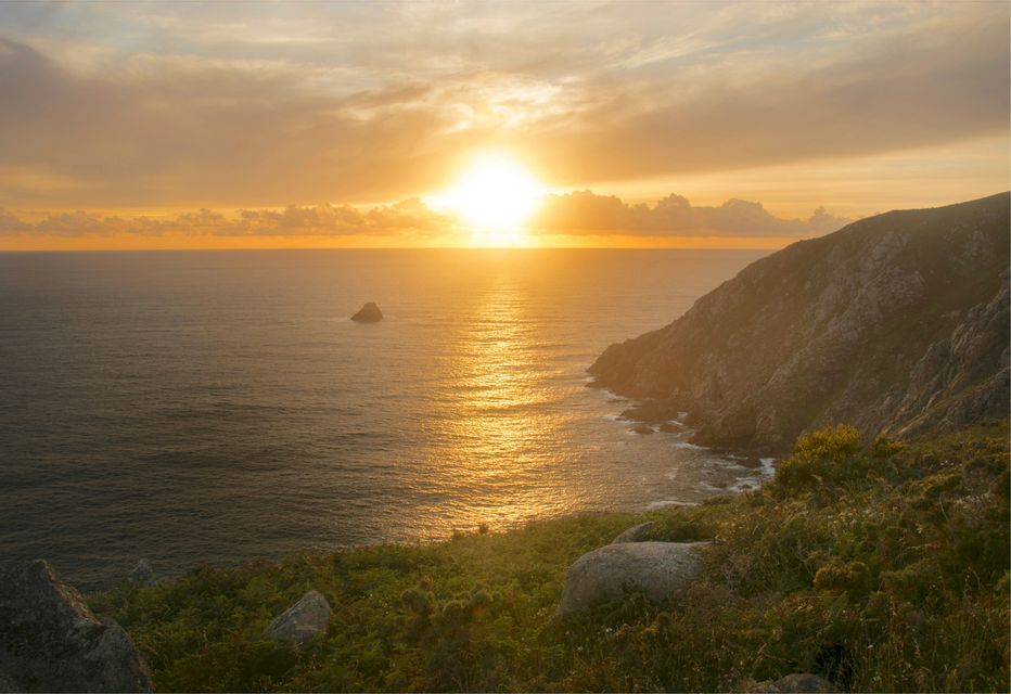 El sol se pone sobre el océano, su luz dorada se refleja en el agua junto a un acantilado rocoso y verde.
