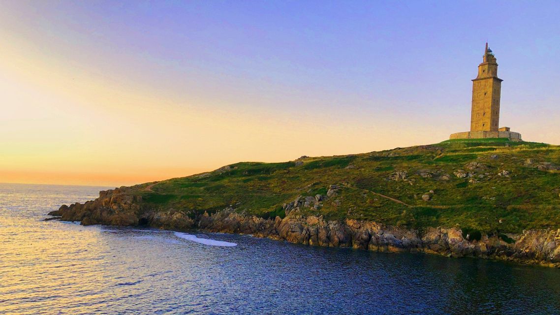 Un faro de piedra se asienta en una colina cubierta de hierba con vistas a una costa rocosa y un mar en calma al atardecer.