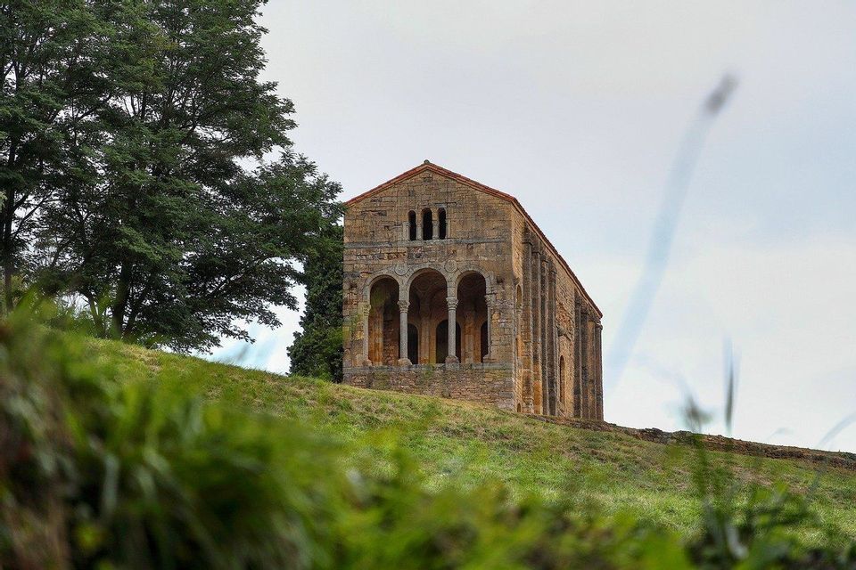 Un antiguo edificio de piedra con un pórtico abovedado se alza en una colina cubierta de hierba junto a un árbol, visto desde un ángulo bajo.