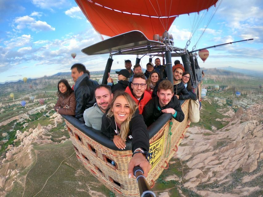 Un gruppo WeRoad si scatta un selfie all'interno di una cesta di mongolfiera che sorvola una valle rocciosa con altre mongolfiere nel cielo.