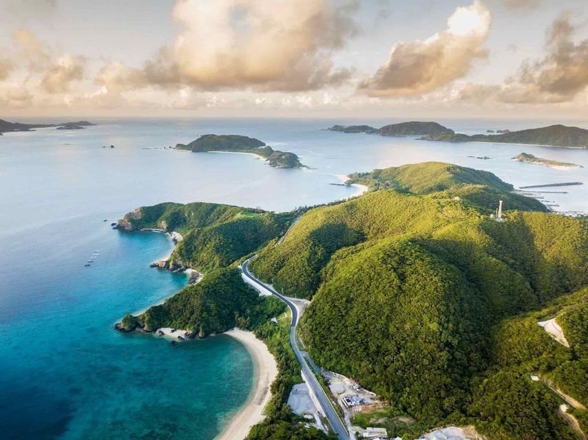 Vista aérea de una carretera sinuosa a lo largo de una costa de exuberantes colinas verdes, con agua turquesa y más islas en la distancia.