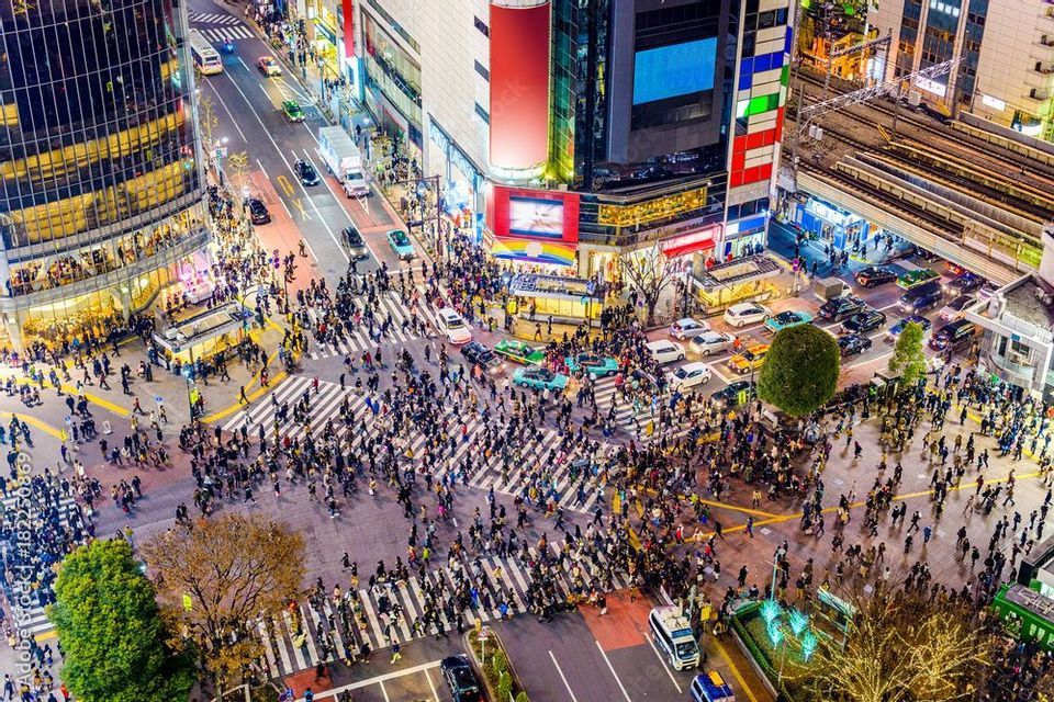 Una vista aérea de una concurrida intersección urbana por la noche, con multitudes cruzando pasos de peatones multidireccionales entre el tráfico.