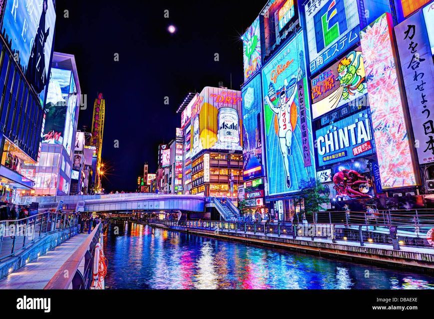 Enormes y coloridos letreros de neón en edificios de la ciudad iluminan un ancho río por la noche mientras la gente pasea por un malecón.