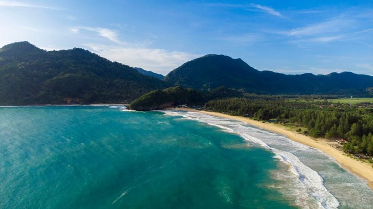 Una vista aerea di una spiaggia curva e sabbiosa con onde turchesi, delimitata da una fitta foresta verde e montagne sotto un cielo blu.