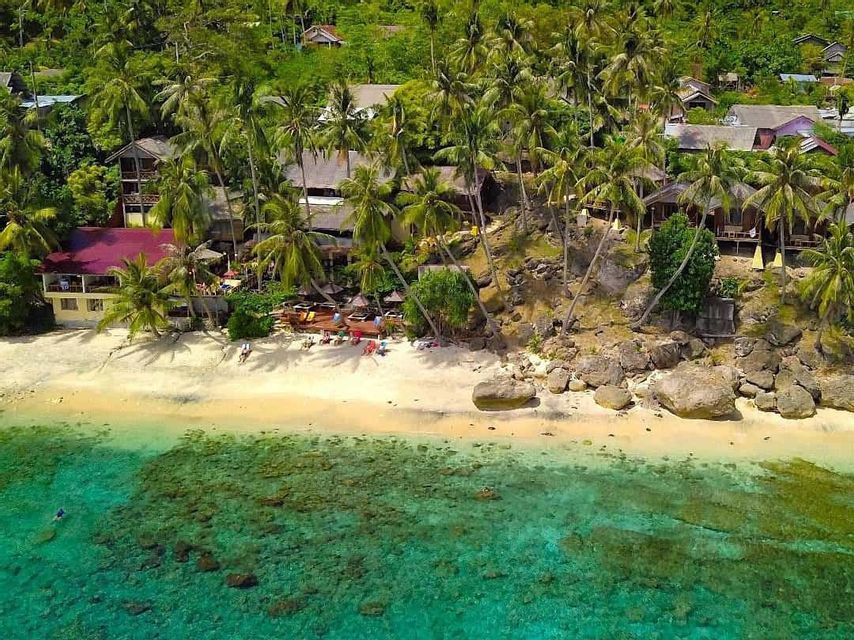 Vista aerea di una spiaggia tropicale con acqua turchese cristallina, una costa sabbiosa ed edifici immersi tra palme su una collina verde.
