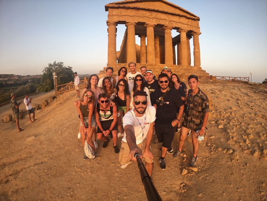 A WeRoad group trip takes a selfie with a selfie stick in front of an ancient Greek temple on a dusty hill at sunset.