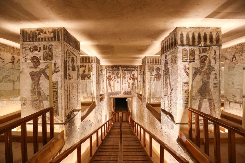 A wooden walkway with railings descends between pillars covered in colorful hieroglyphics inside an ancient Egyptian tomb.