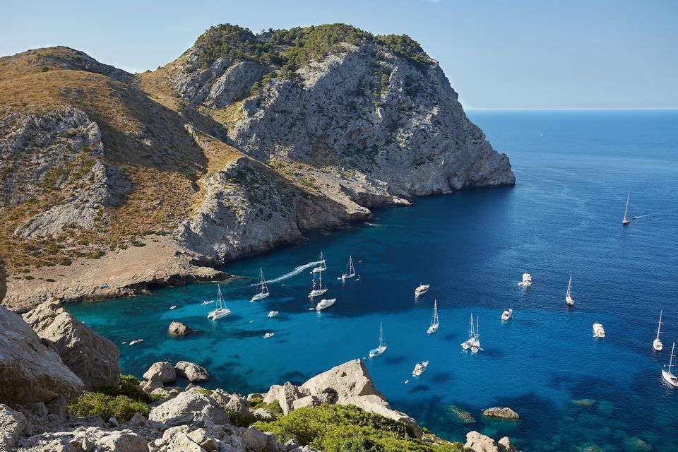 Vista aerea di una caletta rocciosa con acqua turchese, dove numerose barche a vela e yacht sono ormeggiati in una giornata limpida.