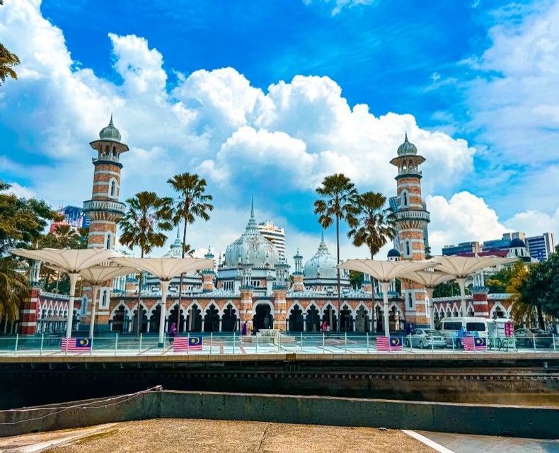Una moschea sontuosa con minareti e cupole a strisce si erge oltre un fiume, con palme e grandi ombrelloni bianchi in primo piano.