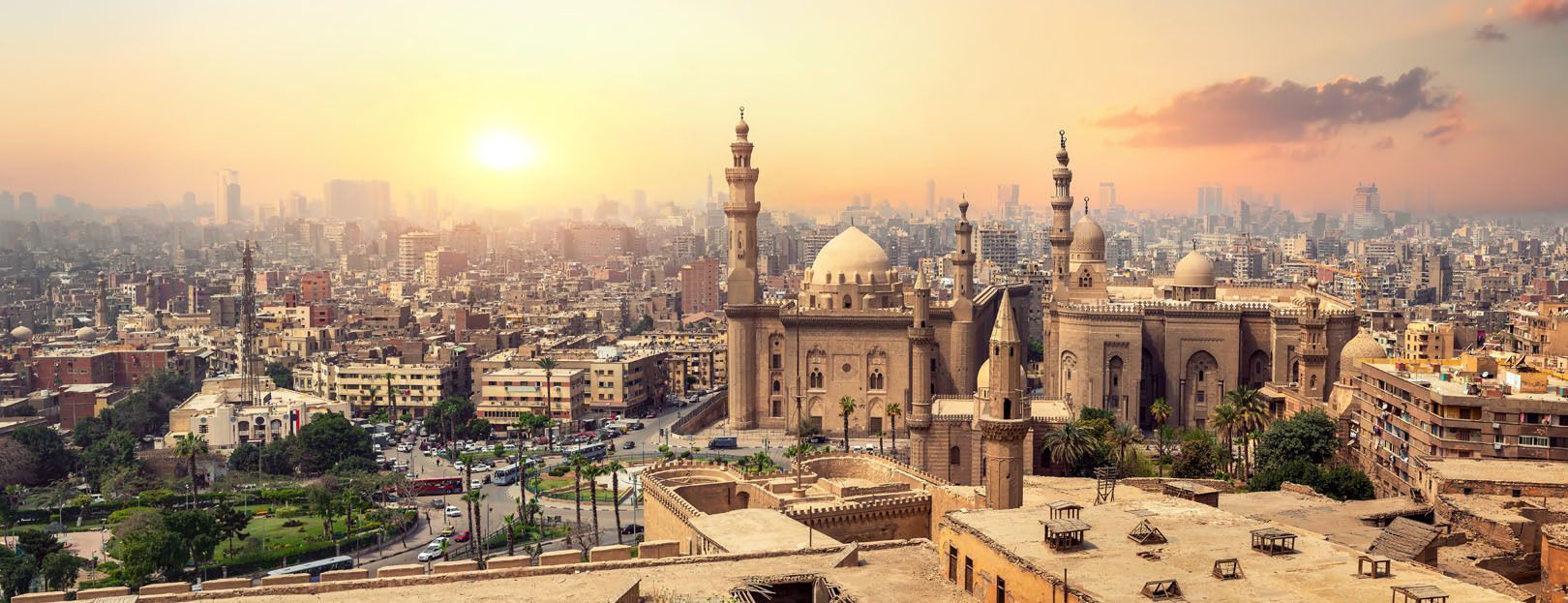 A panoramic view of a large mosque with domes and minarets overlooking a dense cityscape under a hazy, golden sunset.