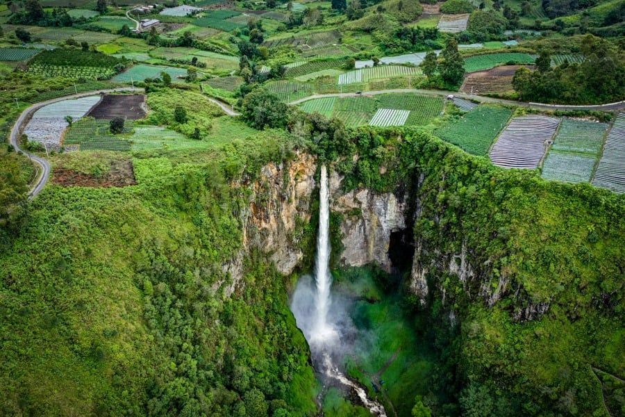 Vista aerea di un'imponente cascata che si getta in una gola verdeggiante, circondata da un mosaico di campi coltivati verdi.