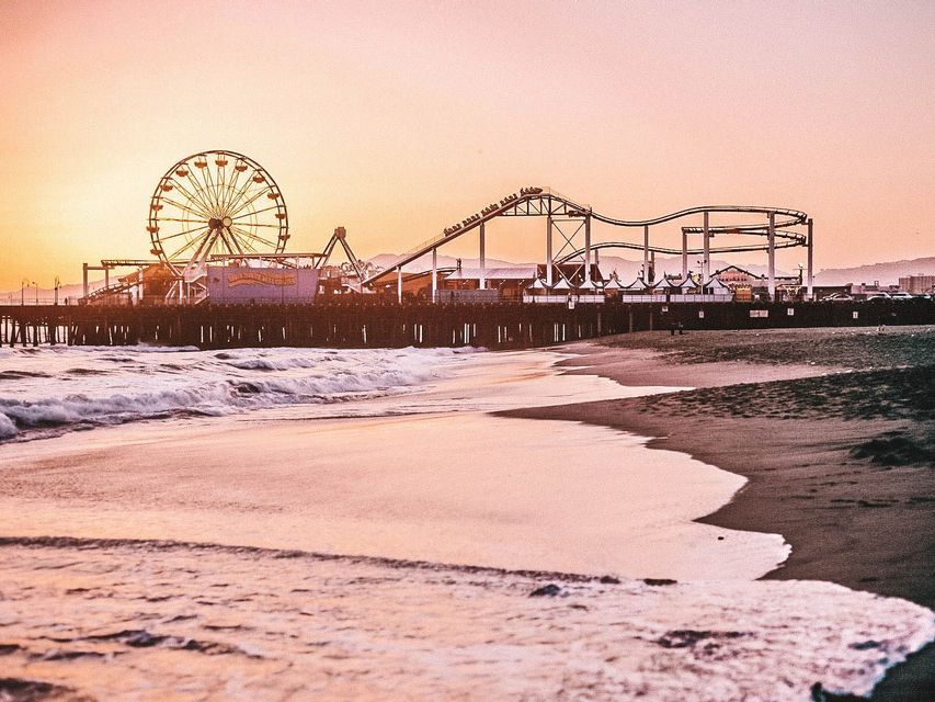 An amusement park pier with a Ferris wheel and roller coaster on a beach, viewed as waves wash ashore during sunset.