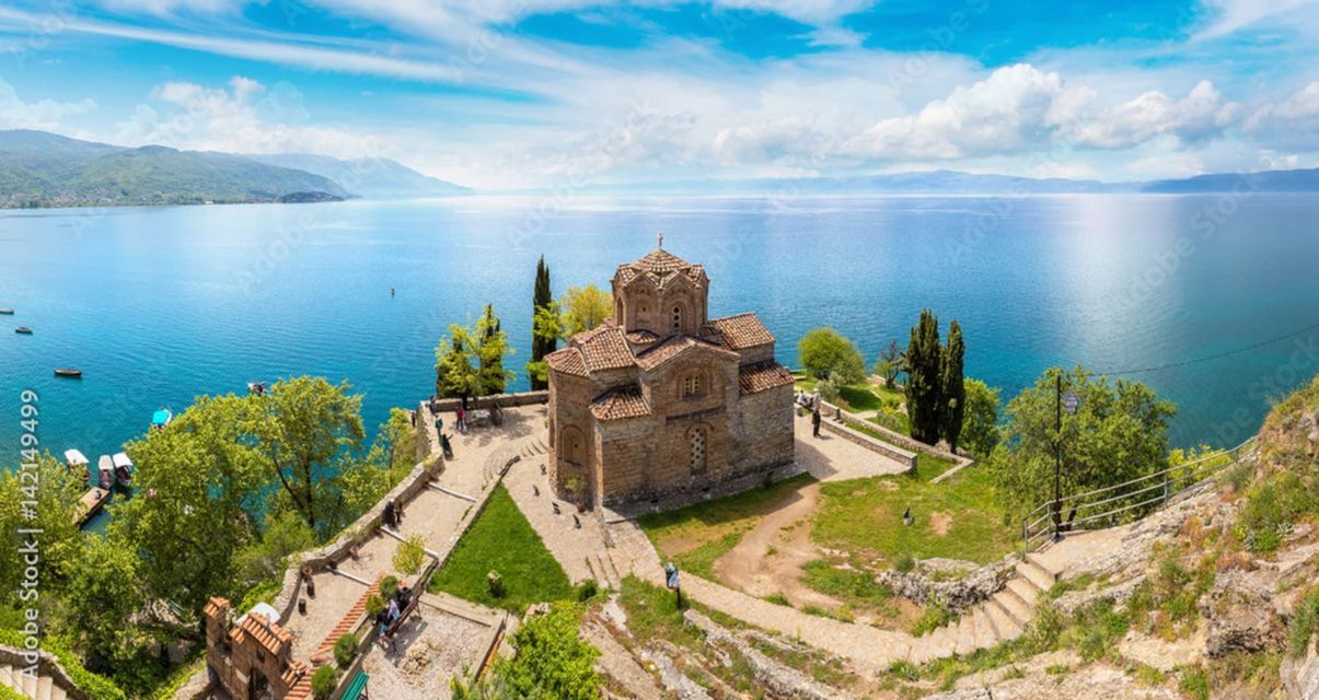 Una vista panorámica de una antigua iglesia de piedra en un acantilado con vistas a un vasto lago azul, con montañas distantes bajo un cielo nublado.