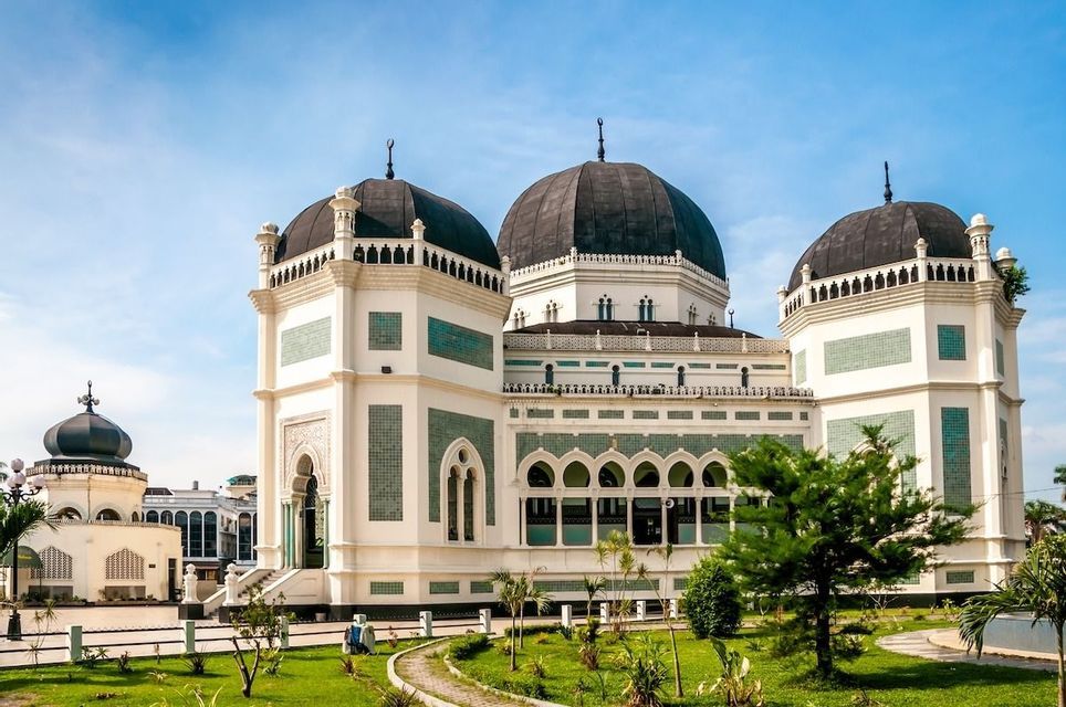 Una moschea bianca ornata con grandi cupole nere e pareti di piastrelle verdi, circondata da un giardino lussureggiante sotto un cielo azzurro chiaro.
