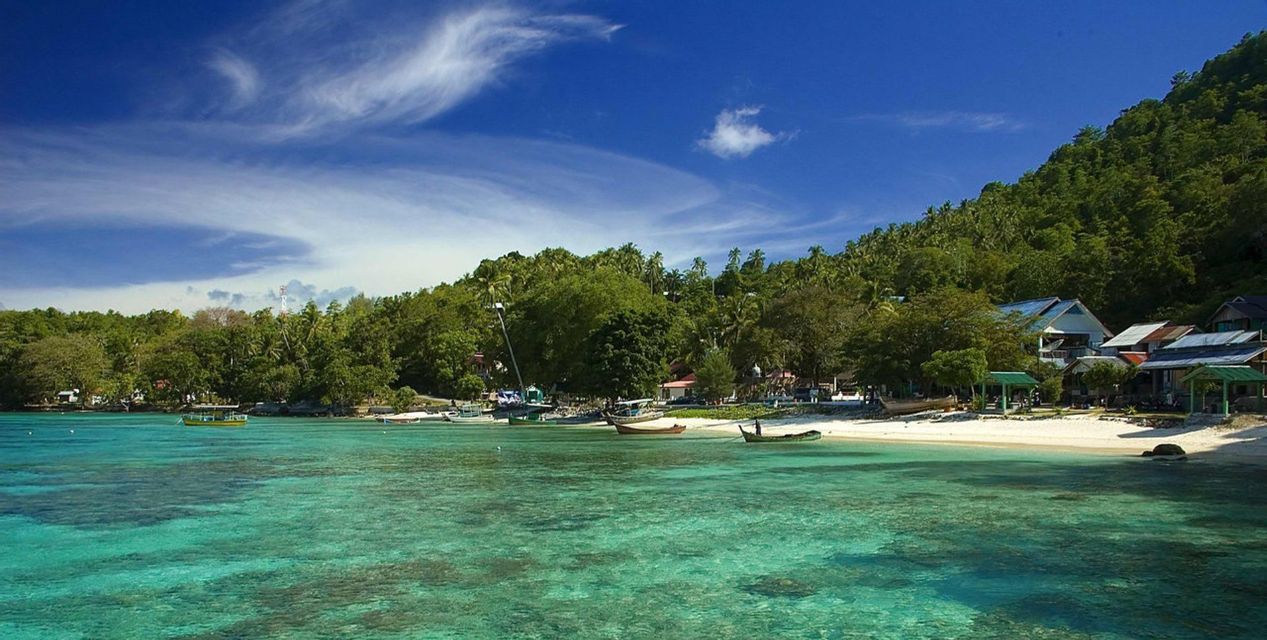 Una vista di una baia tropicale con acqua turchese cristallina, una spiaggia di sabbia bianca con barche e un villaggio immerso in una collina boscosa.